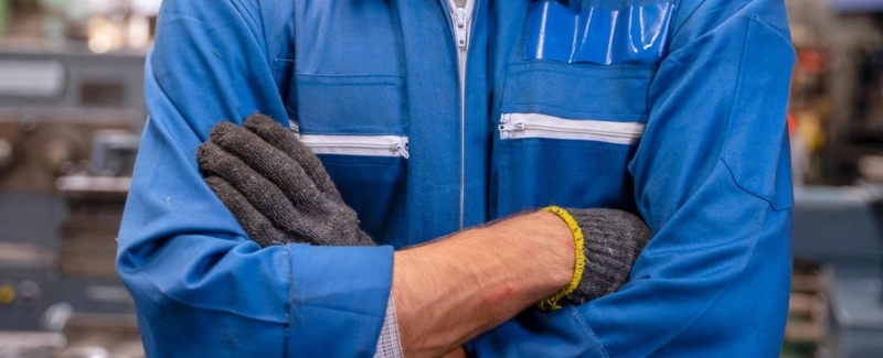 Operario industrial con mono azul y guantes de seguridad cruzado de brazos en un taller técnico, representando gestión y dirección de mantenimiento